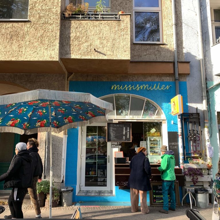 front view of the rudimarie cafe, blue walls and a big window, a big umbrella and 4 people in front