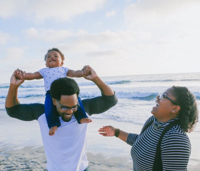 dad with a baby on his neck and his wife on the beach